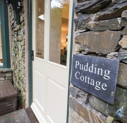 An exterior view of a stone cottage wall with a sign reading Pudding Cottage and a white door at Pudding Cottage in Ambleside