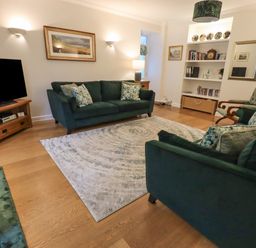 A living room with two green sofas a wooden tv stand with a television a white rug wooden floor shelves with books and decorative plates at Pudding Cottage Ambleside