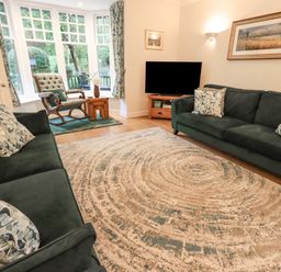 A living room with two sofas, a rocking chair by a bay window, a large rug, a television on a wooden stand, and framed artwork on the walls at Pudding Cottage in Ambleside