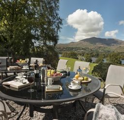 An outdoor patio with round glass tables set with wine glasses cups books fruit and bottles with chairs around overlooking hills and a lake at Tent Lodge in Coniston