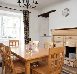 A dining room with a wooden table and chairs a bottle of wine three wine glasses a window with patterned curtains a wall clock and a cream colored stove at Pollys Cottage in Langdale
