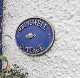 A blue oval sign with gold letters reading Springwell Cottage on a white textured wall with plants nearby at Springwell Cottage Ambleside