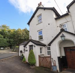 The exterior of a white cottage with a wooden door a small gate potted plants and signage on a paved street at Gale Lodge Cottage in Ambleside