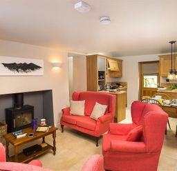 A living room with red upholstered armchairs and a sofa around a wood burning stove with a wooden coffee table and a dining area with wooden chairs at Barn Cottage in Bassenthwaite near Keswick