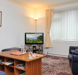 A living room with a wooden coffee table, glasses, a television on a stand, a floor lamp, a leather sofa, and a window with vertical blinds at Mosslea in Bowness