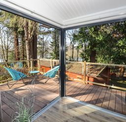 A balcony with two blue chairs and a small table overlooking trees and wooden buildings at Windermere Lodge in Bowness-On-Windermere