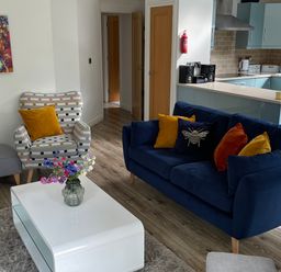 A living room with a blue sofa and colorful cushions a patterned armchair a white coffee table with flowers and an open kitchen at Windermere Lodge in Bowness-On-Windermere