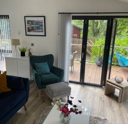 A living room with a blue sofa and armchair a white cabinet and glass doors leading to a wooden deck at Windermere Lodge in Bowness-On-Windermere