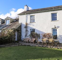 A white house with wooden outdoor chairs and table on a gravel area and a green lawn in front at Robin Cottage in Troutbeck