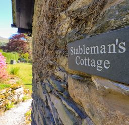 A stone wall with a Stablemans Cottage sign and a garden with plants and trees in the background at Stablemans Cottage at Stepping Stones in Ambleside