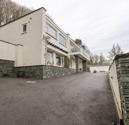 A driveway leading up to a two-story house with stone and white exterior walls with trees in the background at Tanna Hill in Bowness