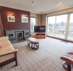 A living room with a sofa, armchair, coffee table, fireplace, television, and large sliding glass doors at Tanna Hill in Bowness