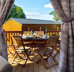 An outdoor wooden dining table with chairs on a deck with food and drinks visible through open curtains at Lancashire Rose in Ambleside