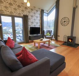 A living room with a grey sofa with red cushions a wooden coffee table a grey armchair a wall clock and a wood burning stove at Idle Mill 2 in Ambleside