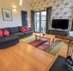 A living room with a grey sofa and red cushions a wooden coffee table on a striped rug a television on a stand floral wallpapered wall and large windows at Idle Mill 2 in Ambleside