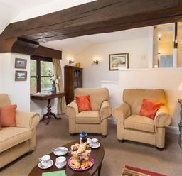 A living room with beige sofas and armchairs a wooden coffee table with tea sets and pastries a stone fireplace and wooden beam at Thirlmere Cottage in Grasmere