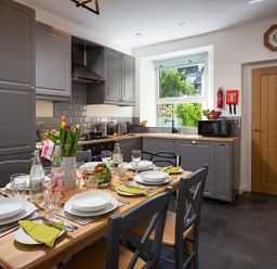 A kitchen with grey cabinets a wooden dining table set with plates glasses and cutlery at Fell Cottage Ambleside