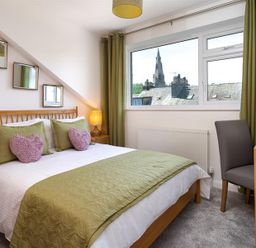 A bedroom with a double bed with green and pink pillows a bedside table with lamp a dressing table with a chair and a window showing rooftops and a church spire Fell Cottage Ambleside