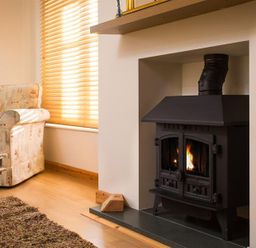 A room with a wood burning stove built into a wall next to a patterned armchair and wooden coffee table at Agathi in Keswick