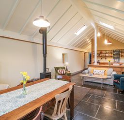 An open living space with dining table flowers wood stove armchairs sofa and kitchen shelves at Old Cary Cottage Henley near Langport