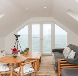 A living room with wooden furniture and a telescope by large windows overlooking the sea at St Julien in Dawlish