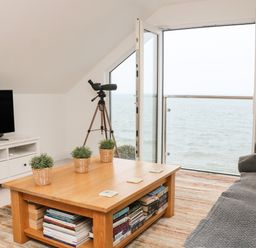 A living room with a gray couch wooden coffee table with plants and books a television on a white stand and a telescope by a window overlooking the sea at St Julien in Dawlish