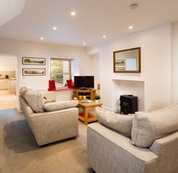 A living room with two beige sofas a wooden coffee table a television on a stand a window with red cushions and a doorway to a kitchen at Fern Cottage in Ambleside