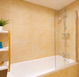 A bathroom with beige tiled walls a white bathtub a glass shower screen and a wooden shelf with a green plant and toiletries at Fern Cottage in Ambleside