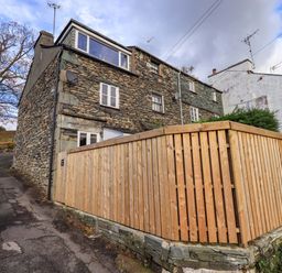 A stone house with white windows and a wooden fence on a sloped street at Penny Red Cottage in Ambleside