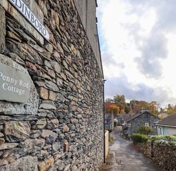 A stone wall with Penny Red Cottage sign along a narrow street with houses and trees in Ambleside