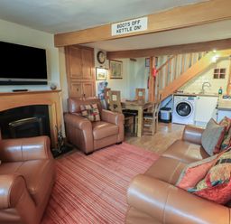 A living room with brown leather sofas and armchairs a wooden dining table under stairs an open plan kitchen with a washing machine and a wall mounted television at Heron Cottage in Ambleside