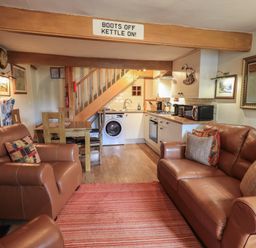 A living room with leather sofas a wooden dining table and chairs a kitchen with appliances under stairs at Heron Cottage in Ambleside