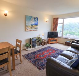 A living room with a wooden dining table and chairs a leather sofa and armchair a colorful rug a tv on a wooden stand and large window at Fellcroft in Troutbeck