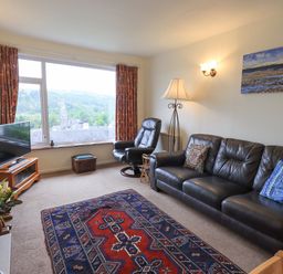 A living room with a leather sofa two armchairs a patterned rug a television and a large window showing a village view at Fellcroft in Troutbeck