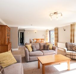 A living room with sofas, wooden coffee table, dining table with chairs, wooden cabinet, and windows with curtains at Ghyll View in Ambleside
