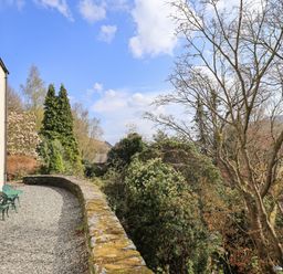 A gravel patio with green metal chairs and a stone wall next to a house with trees and shrubs in the background at Rawfell in Great Langdale