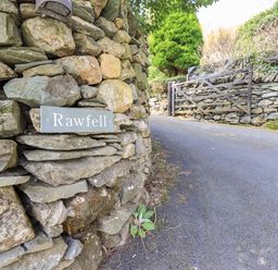 A stone wall with a Rawfell sign next to a paved road and a wooden gate at Rawfell Great Langdale