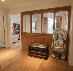 A hallway with wooden floor a black chest and a folded baby high chair by wooden window panels looking into a sitting room at Orchard House in Ambleside