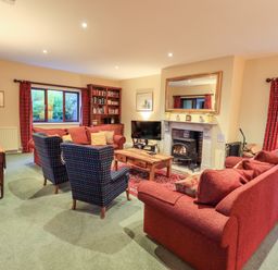 A living room with red sofas, two patterned armchairs, a wooden coffee table, a fireplace, a TV, a bookcase, and windows with red curtains at Low House in Ullswater