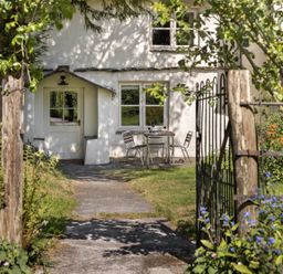 A pathway leading to a white cottage entrance with a table and two chairs outside surrounded by plants and flowers at High Lindeth in Bowness