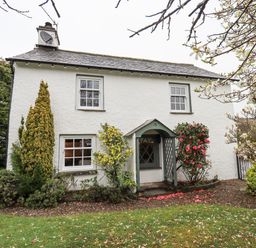 A white house with a slate roof and a small porch surrounded by shrubs and trees at Syke Cottage in Hawkshead