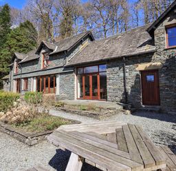 Outside view of a stone house with wooden windows and doors surrounded by trees and garden beds with a wooden picnic table in the foreground at The Stables in Ambleside