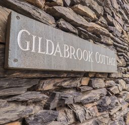 A stone wall with a rectangular sign that reads Gildabrook Cottage in Bowness