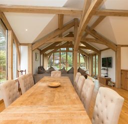 A dining room with a long wooden table and beige upholstered chairs near wooden beams and large windows at Old Droomer in Windermere