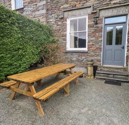 An outdoor area with a wooden picnic table and benches beside a stone building with a grey door and window surrounded by green bushes at Fairy Glen in Grasmere near Ambleside