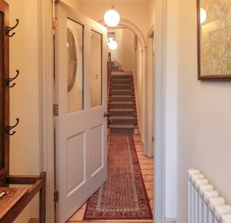 A hallway with a white door open showing a carpet runner on wooden floor leading to stairs with a blue runner and round ceiling lights at Fairy Glen in Grasmere near Ambleside