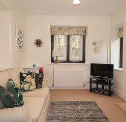 A living room with a beige sofa and floral cushions a TV on a glass stand a window with floral curtains and a fireplace at Larch Cottage in Hawkshead