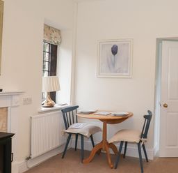 A small dining area with a wooden table, two blue chairs with cushions, a lamp on a windowsill, and framed artwork on the walls at Larch Cottage in Hawkshead