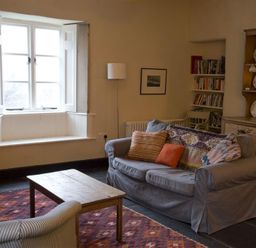 A living room with a wooden coffee table a sofa with cushions an armchair a window a floor lamp a bookcase and a wooden cabinet with plates at Thomas Cottage in Hartsop