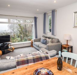 A living room with gray sofas a wooden table a television and large windows showing trees at Helm Crag in Grasmere